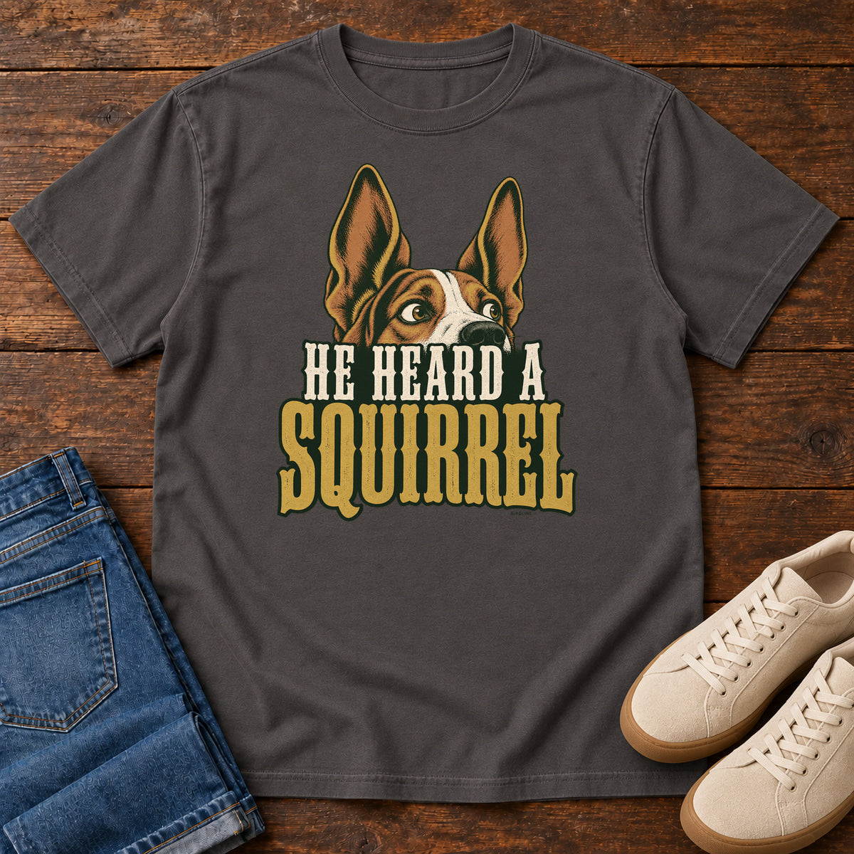 Graphite He Heard a Squirrel T-Shirt with an alert dog with upright ears above He Heard a Squirrel lettering; front flat lay on wood.