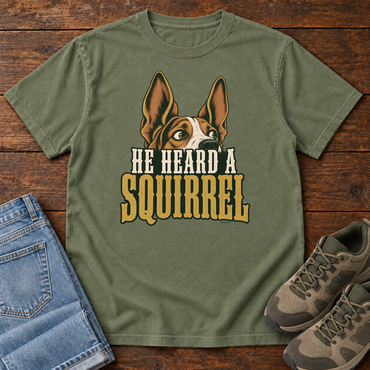 Moss He Heard a Squirrel T-Shirt with an alert dog with upright ears above He Heard a Squirrel lettering; front flat lay on wood.