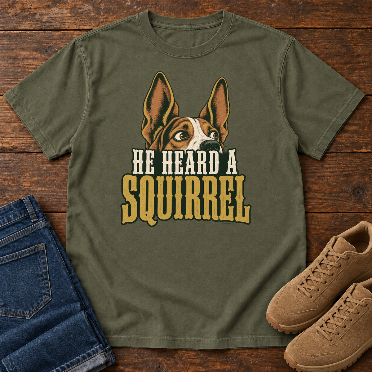 Sage He Heard a Squirrel T-Shirt with an alert dog with upright ears above He Heard a Squirrel lettering; front flat lay on wood.