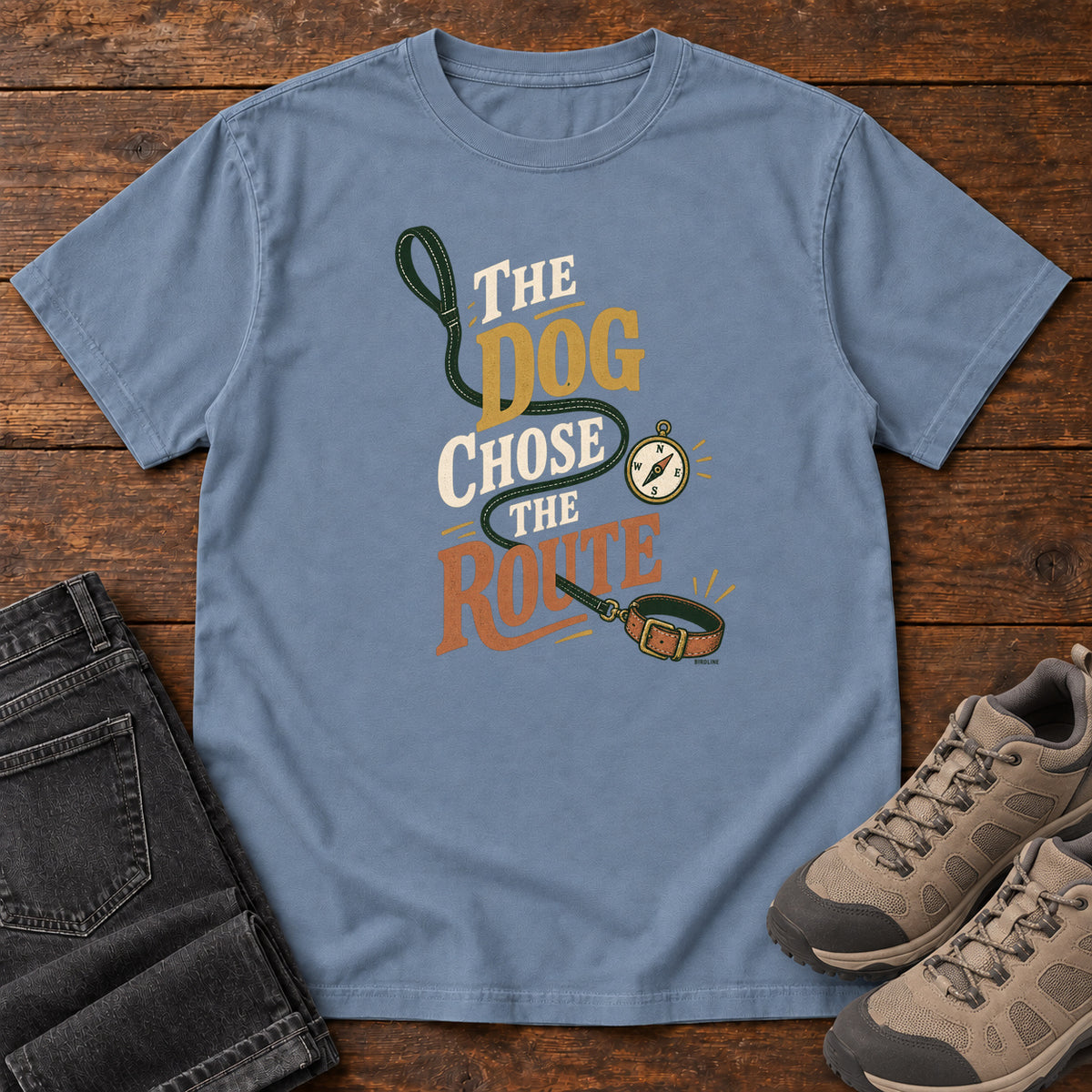 Blue Jean The Dog Chose the Route T-Shirt with a green leash, dog collar and compass around stacked lettering; front flat lay on wood.