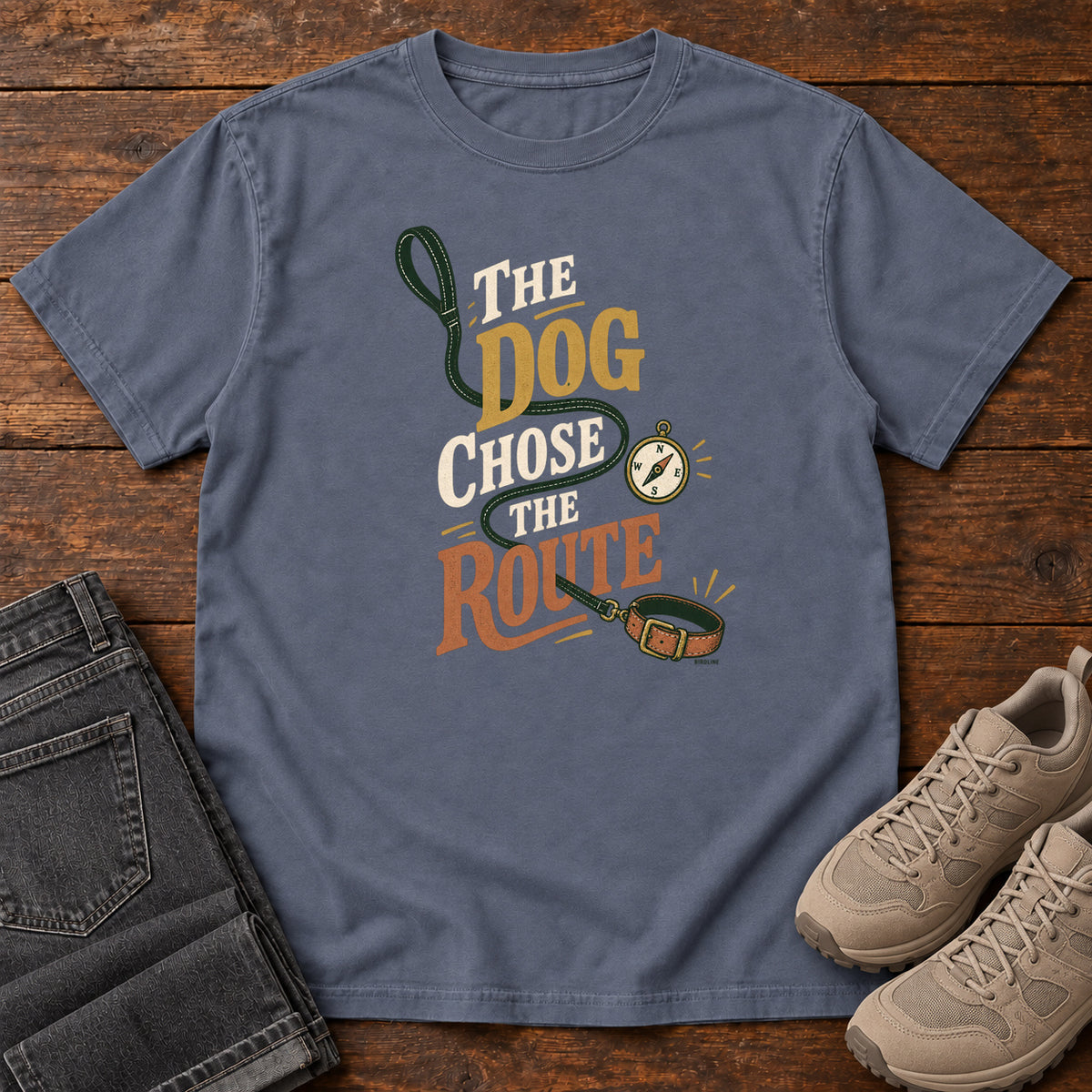Denim The Dog Chose the Route T-Shirt with a green leash, dog collar and compass around stacked lettering; front flat lay on wood.