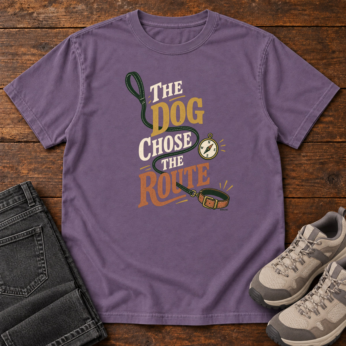 Grape The Dog Chose the Route T-Shirt with a green leash, dog collar and compass around stacked lettering; front flat lay on wood.