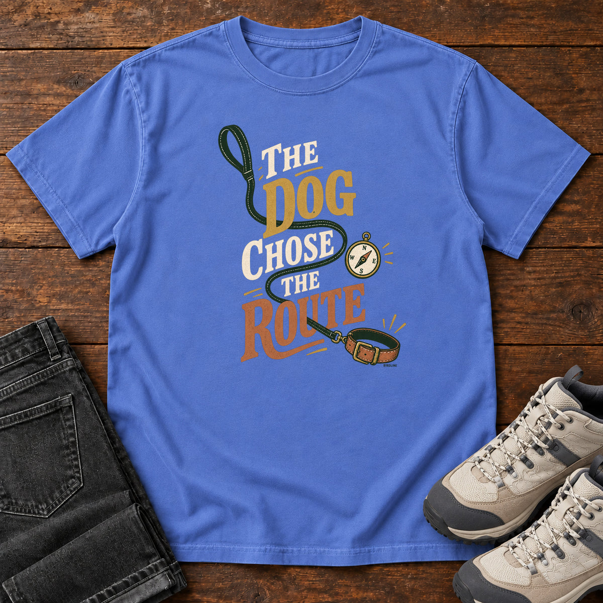 Mystic Blue The Dog Chose the Route T-Shirt with a green leash, dog collar and compass around stacked lettering; front flat lay on wood.