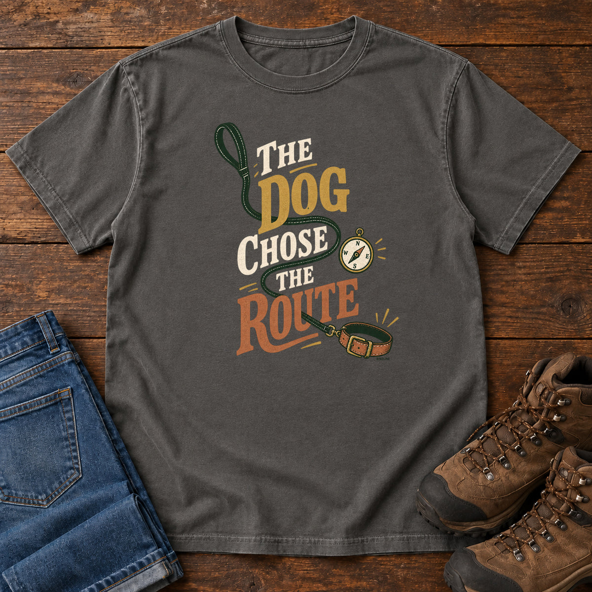 Pepper The Dog Chose the Route T-Shirt with a green leash, dog collar and compass around stacked lettering; front flat lay on wood.