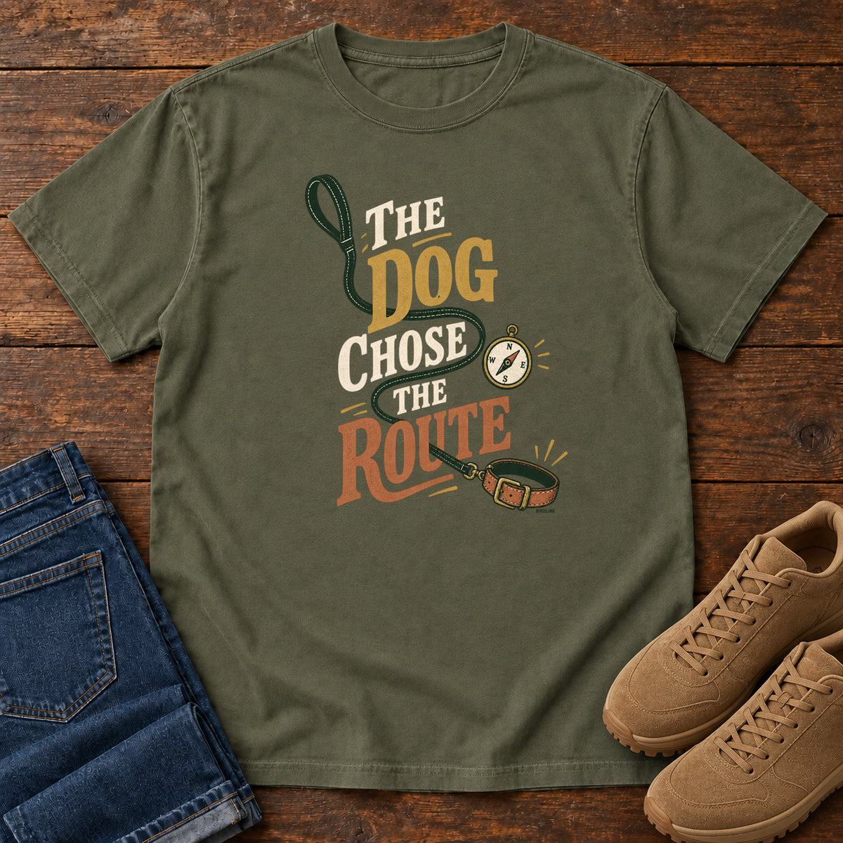 Sage The Dog Chose the Route T-Shirt with a green leash, dog collar and compass around stacked lettering; front flat lay on wood.