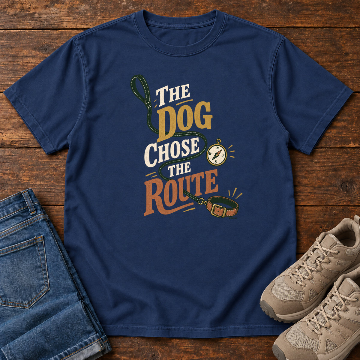 True Navy The Dog Chose the Route T-Shirt with a green leash, dog collar and compass around stacked lettering; front flat lay on wood.
