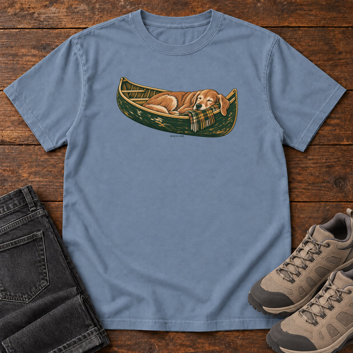 Blue Jean Retriever Canoe Birdline T-Shirt with a golden dog resting in a green canoe, front flat lay on wood.