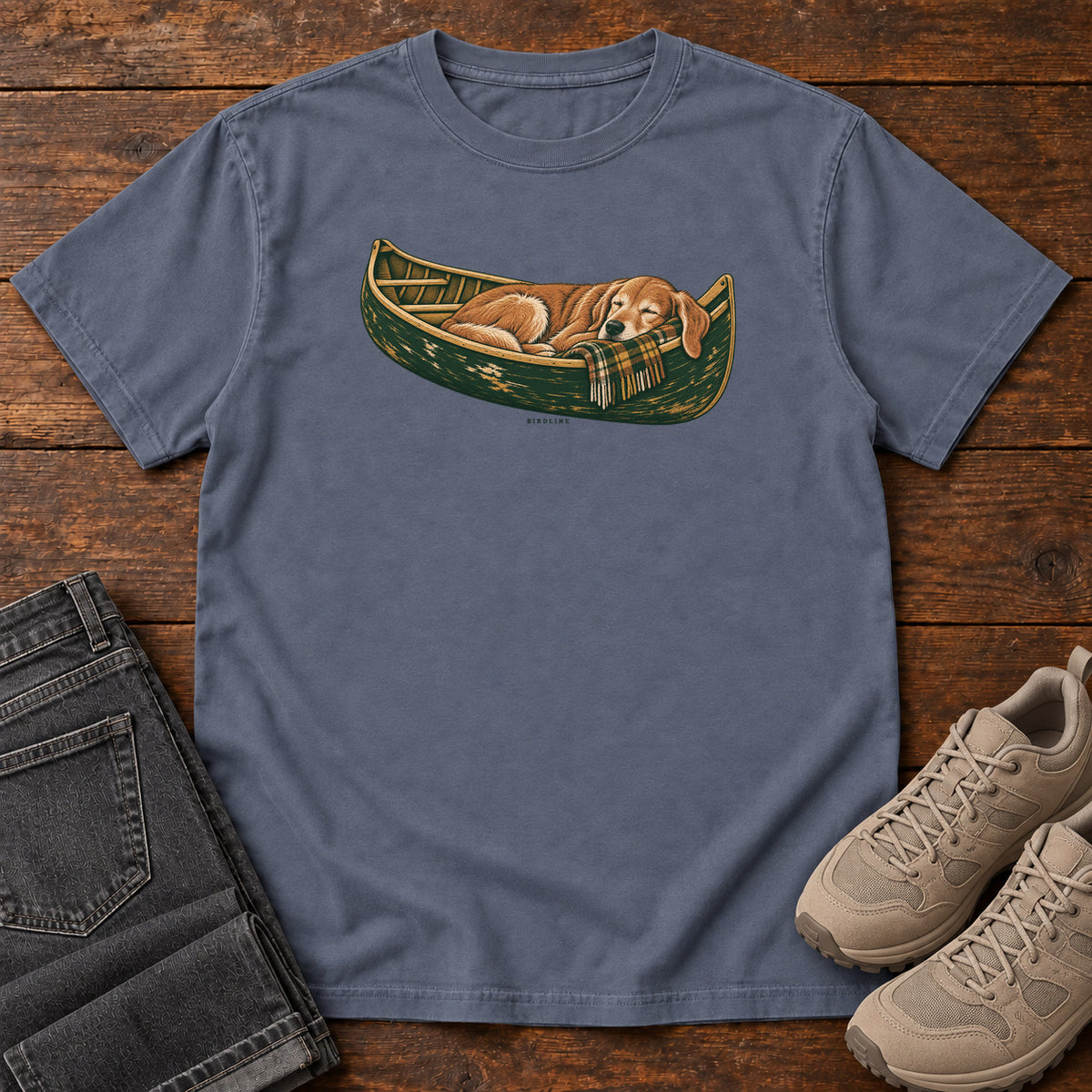 Denim Retriever Canoe Birdline T-Shirt with a golden dog resting in a green canoe, front flat lay on wood.