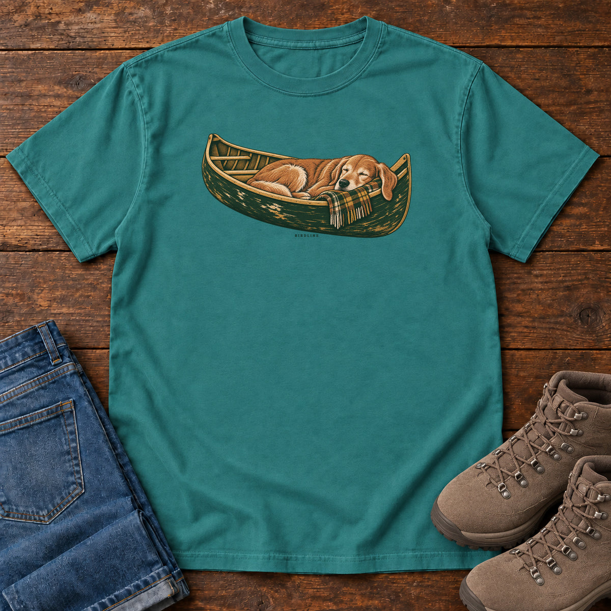 Emerald Retriever Canoe Birdline T-Shirt with a golden dog resting in a green canoe, front flat lay on wood.