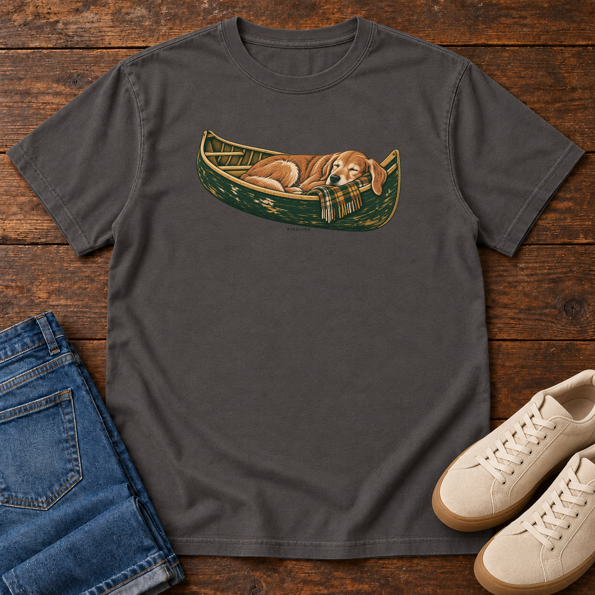 Graphite Retriever Canoe Birdline T-Shirt with a golden dog resting in a green canoe, front flat lay on wood.