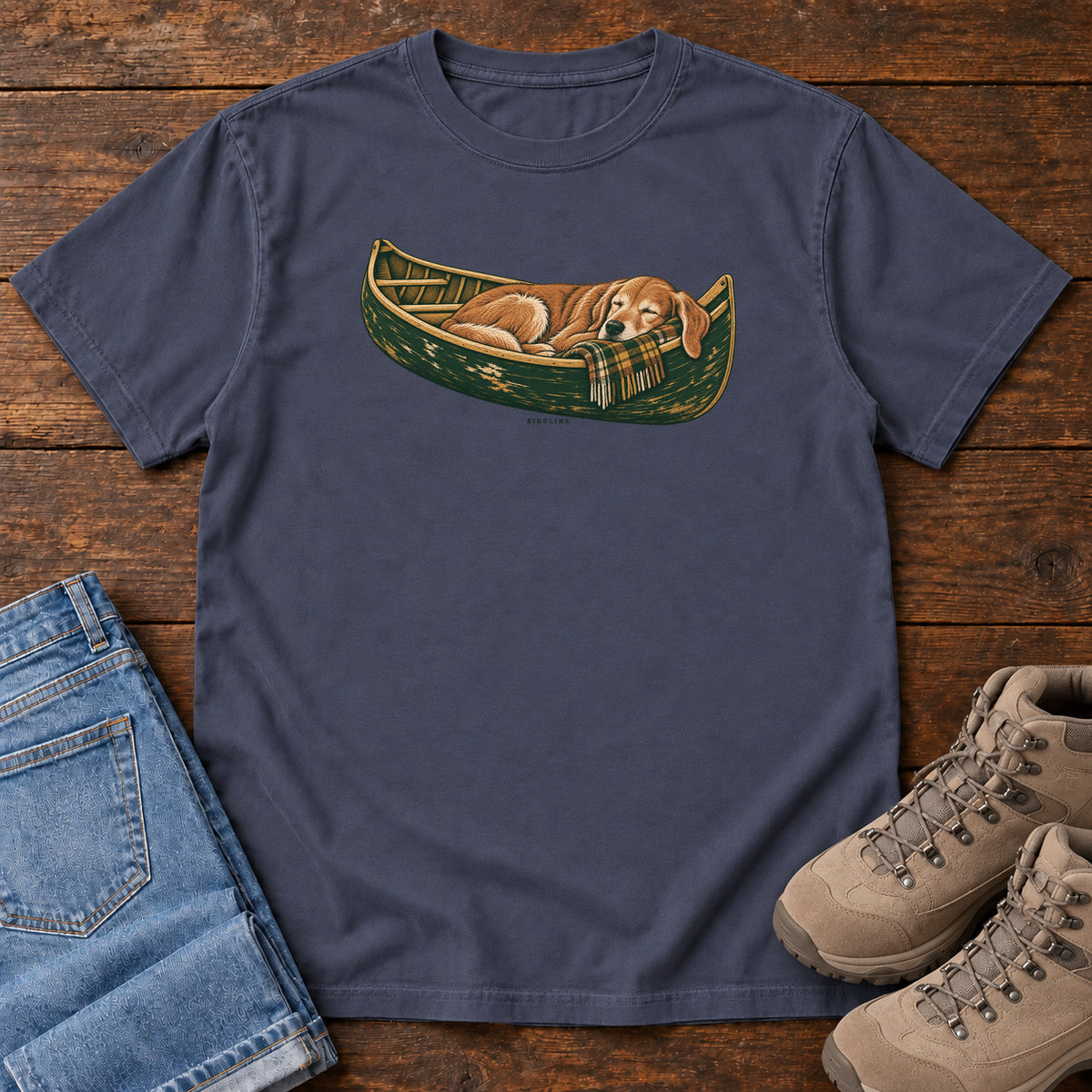Navy Retriever Canoe Birdline T-Shirt with a golden dog resting in a green canoe, front flat lay on wood.