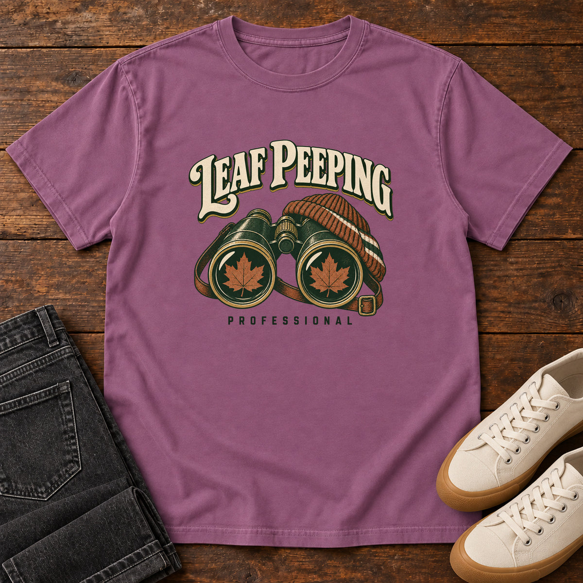 Leaf Peeping Professional T-Shirt in Berry, front laid flat on wood, featuring binoculars with orange leaves in the lenses and a striped knit hat beneath cream lettering.