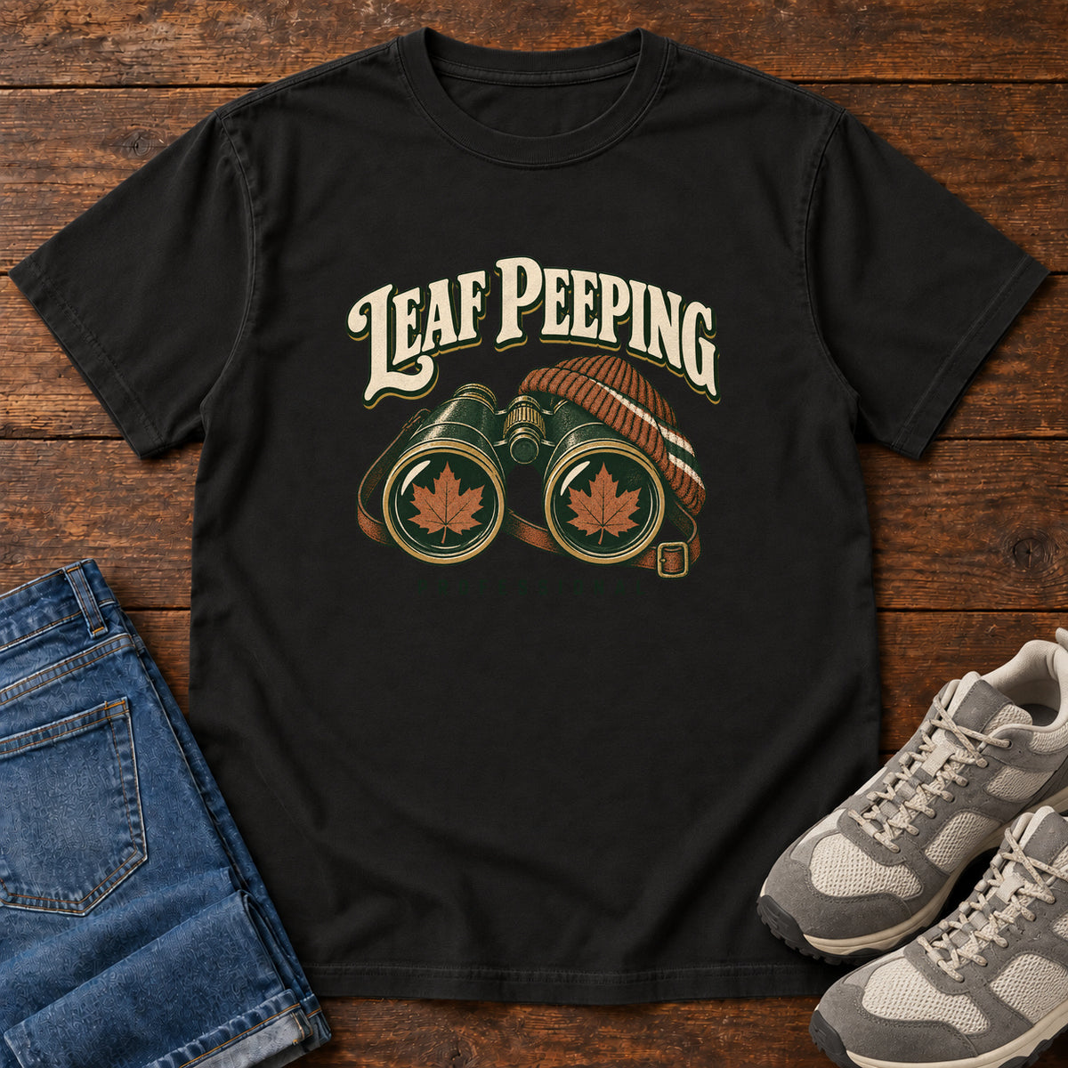 Leaf Peeping Professional T-Shirt in Black, front laid flat on wood, featuring binoculars with orange leaves in the lenses and a striped knit hat beneath cream lettering.