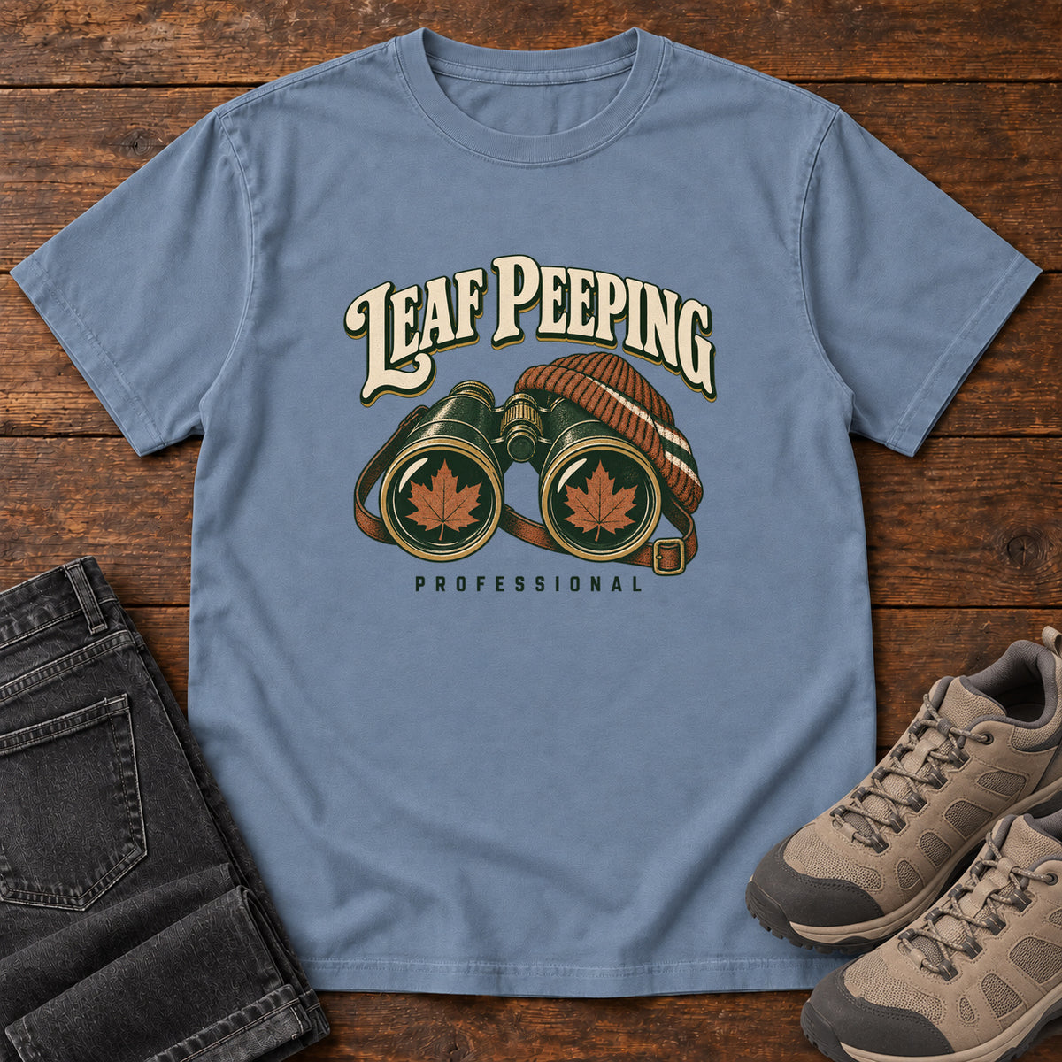 Leaf Peeping Professional T-Shirt in Blue Jean, front laid flat on wood, featuring binoculars with orange leaves in the lenses and a striped knit hat beneath cream lettering.