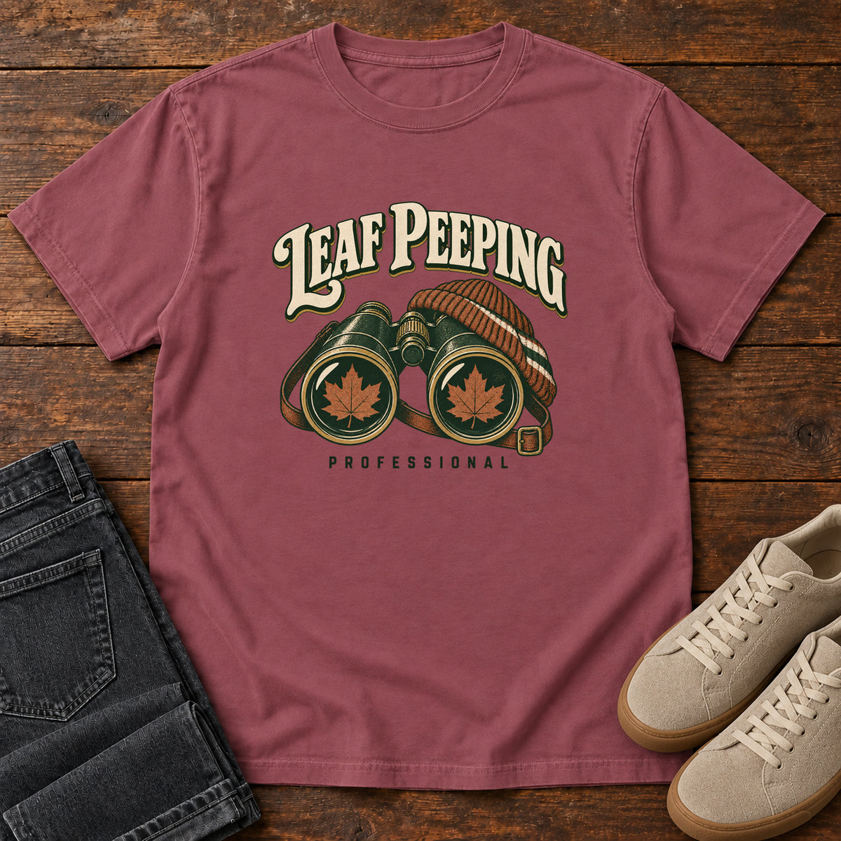 Leaf Peeping Professional T-Shirt in Brick, front laid flat on wood, featuring binoculars with orange leaves in the lenses and a striped knit hat beneath cream lettering.