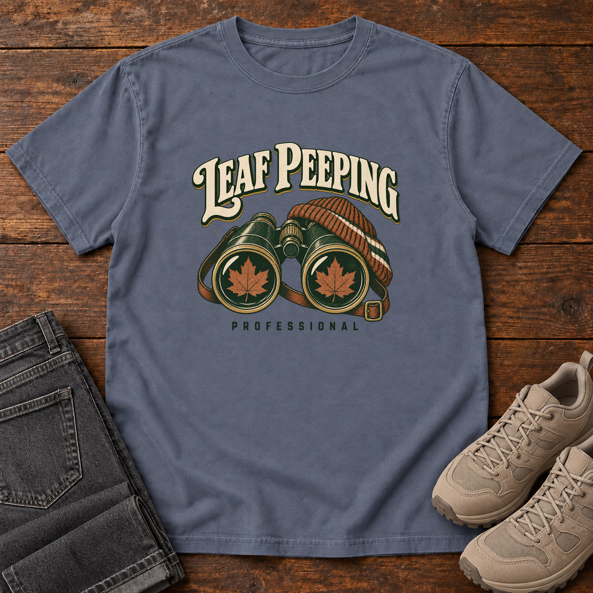Leaf Peeping Professional T-Shirt in Denim, front laid flat on wood, featuring binoculars with orange leaves in the lenses and a striped knit hat beneath cream lettering.