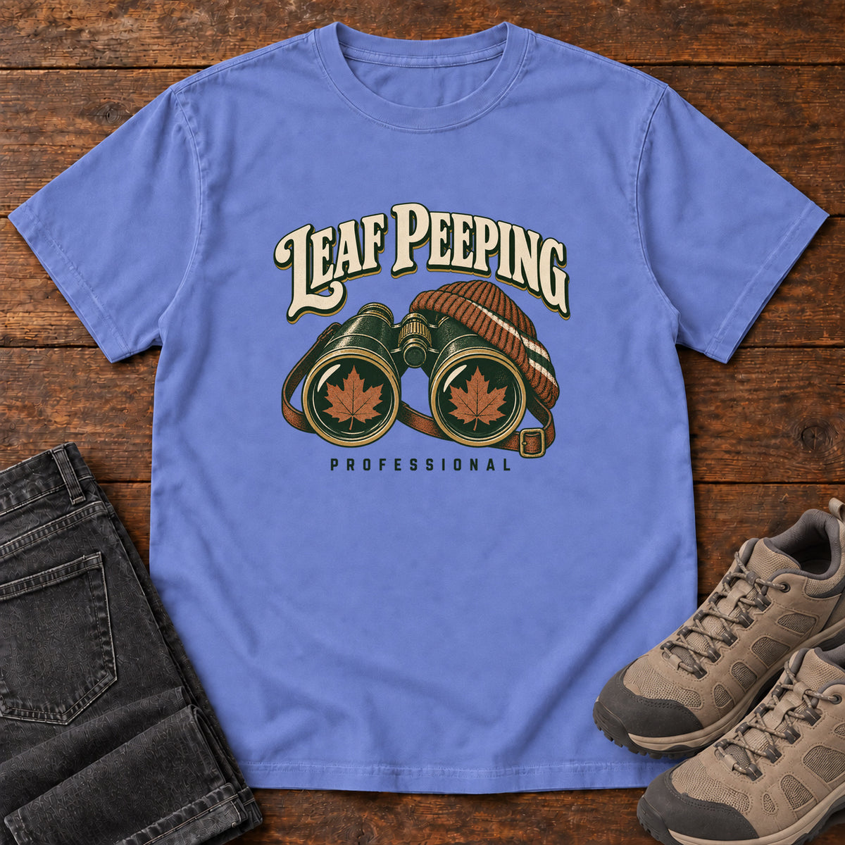 Leaf Peeping Professional T-Shirt in Flo Blue, front laid flat on wood, featuring binoculars with orange leaves in the lenses and a striped knit hat beneath cream lettering.