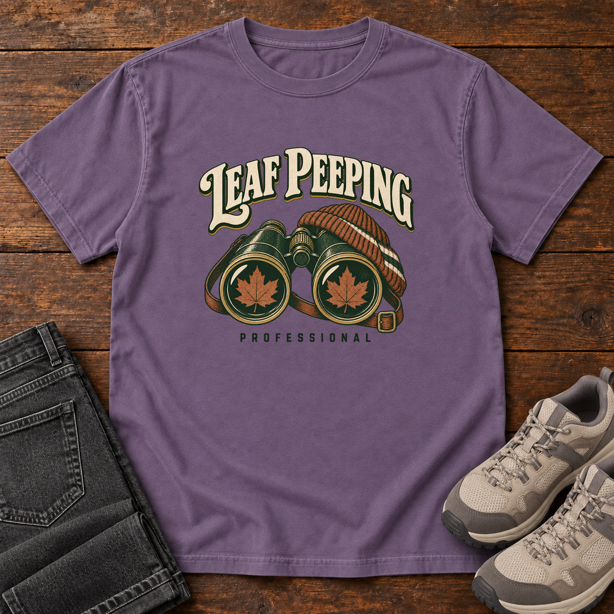 Leaf Peeping Professional T-Shirt in Grape, front laid flat on wood, featuring binoculars with orange leaves in the lenses and a striped knit hat beneath cream lettering.