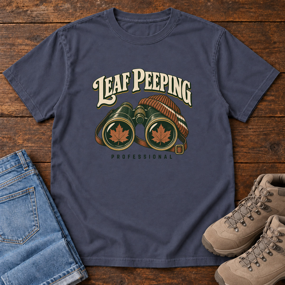 Leaf Peeping Professional T-Shirt in Navy, front laid flat on wood, featuring binoculars with orange leaves in the lenses and a striped knit hat beneath cream lettering.