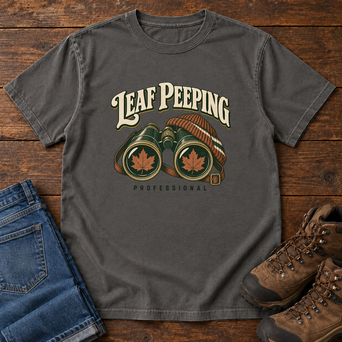 Leaf Peeping Professional T-Shirt in Pepper, front laid flat on wood, featuring binoculars with orange leaves in the lenses and a striped knit hat beneath cream lettering.