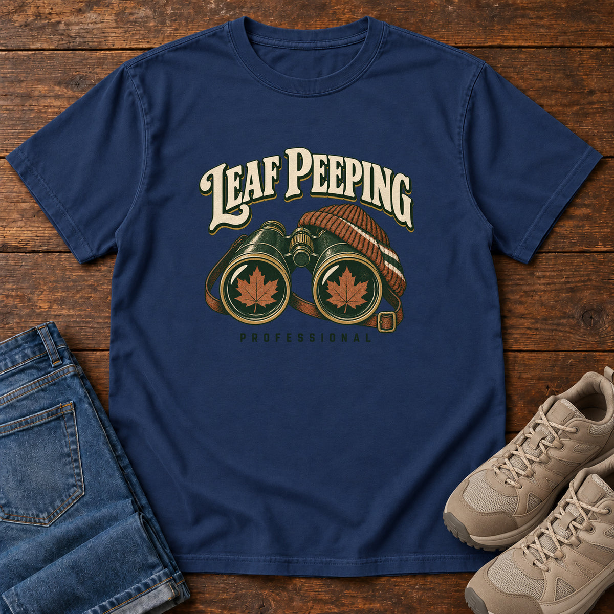 Leaf Peeping Professional T-Shirt in True Navy, front laid flat on wood, featuring binoculars with orange leaves in the lenses and a striped knit hat beneath cream lettering.