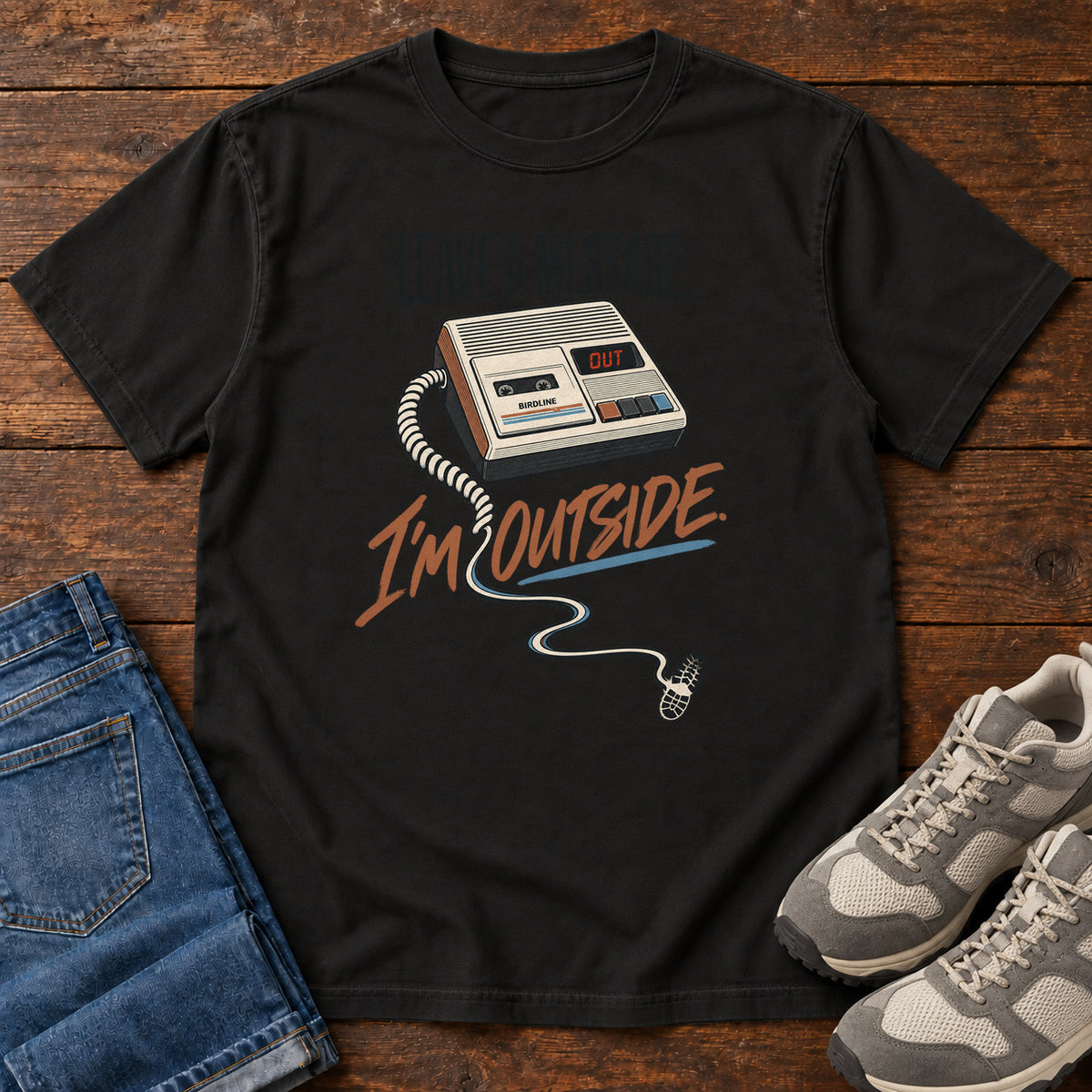 Black Leave a Message I'm Outside T-Shirt with a retro answering machine, trailing cord and orange script, front flat lay on wood.