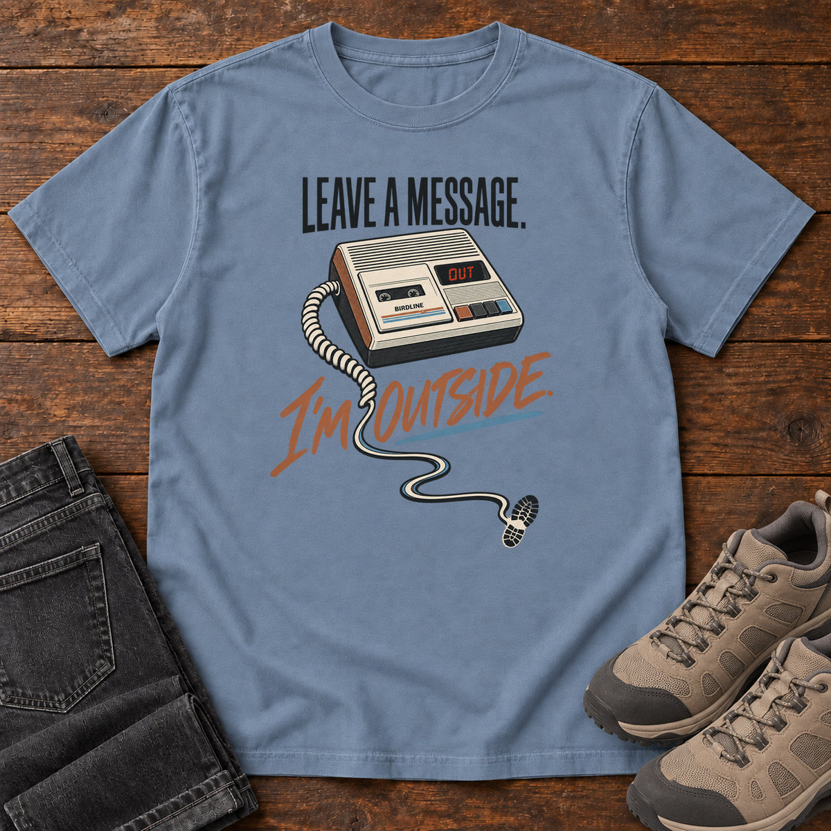 Blue Jean Leave a Message I'm Outside T-Shirt with a retro answering machine, trailing cord and orange script, front flat lay on wood.