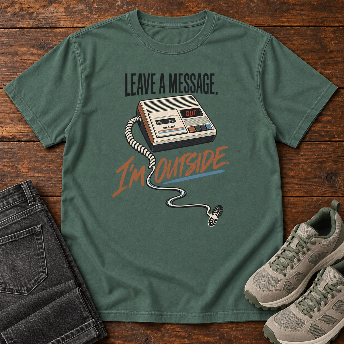 Blue Spruce Leave a Message I'm Outside T-Shirt with a retro answering machine, trailing cord and orange script, front flat lay on wood.