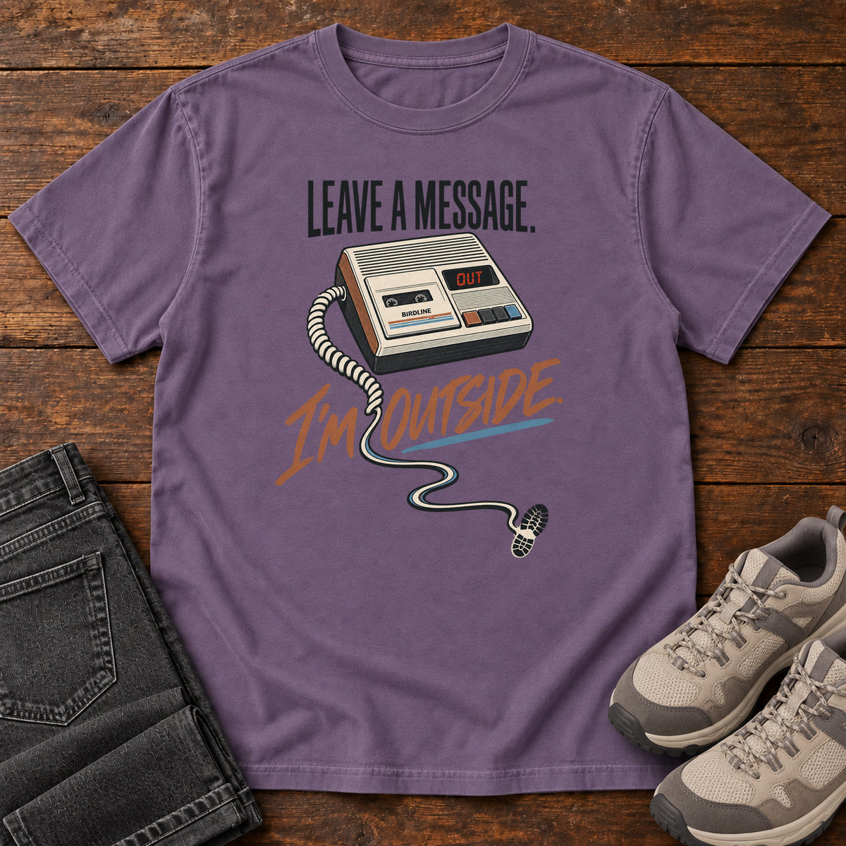 Grape Leave a Message I'm Outside T-Shirt with a retro answering machine, trailing cord and orange script, front flat lay on wood.