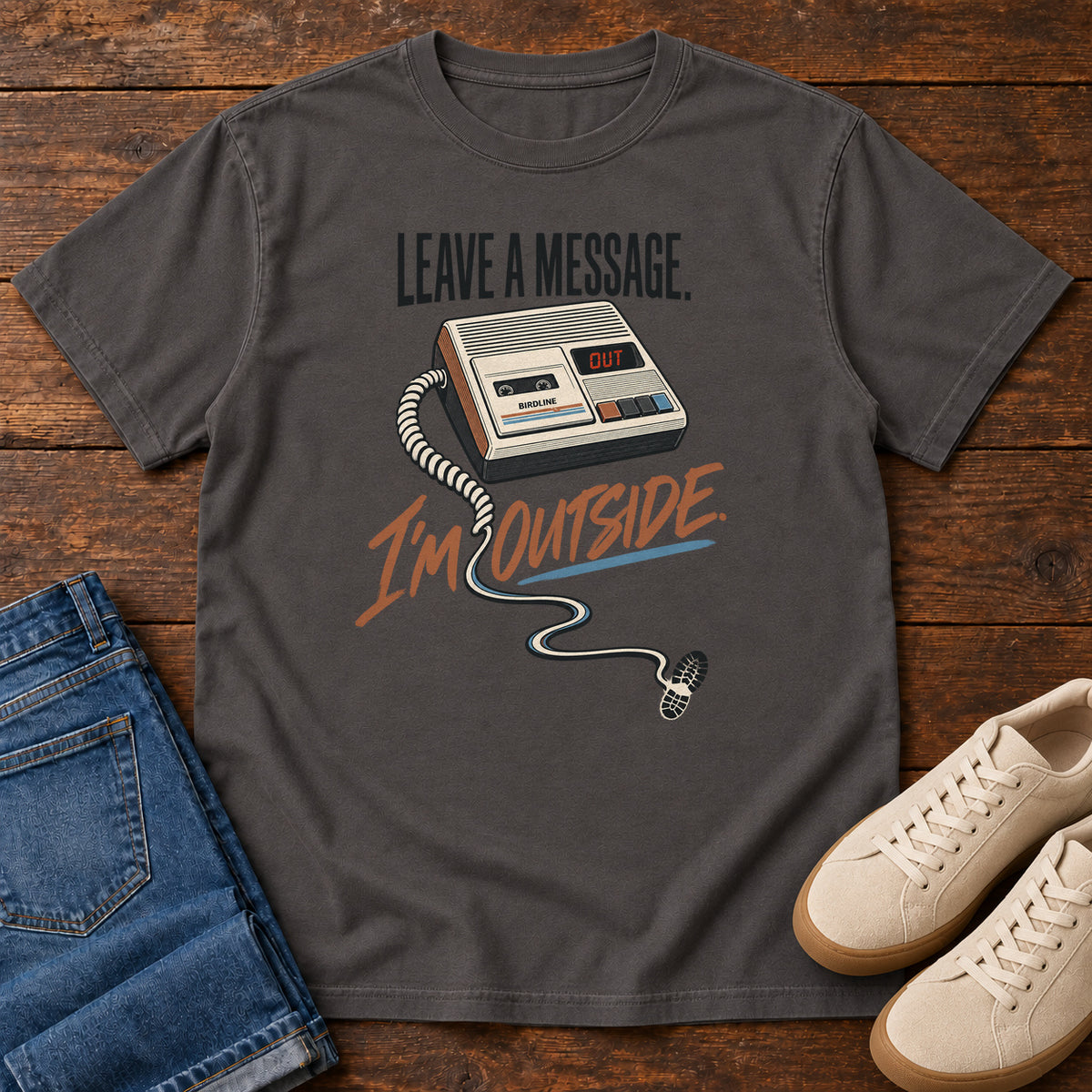 Graphite Leave a Message I'm Outside T-Shirt with a retro answering machine, trailing cord and orange script, front flat lay on wood.