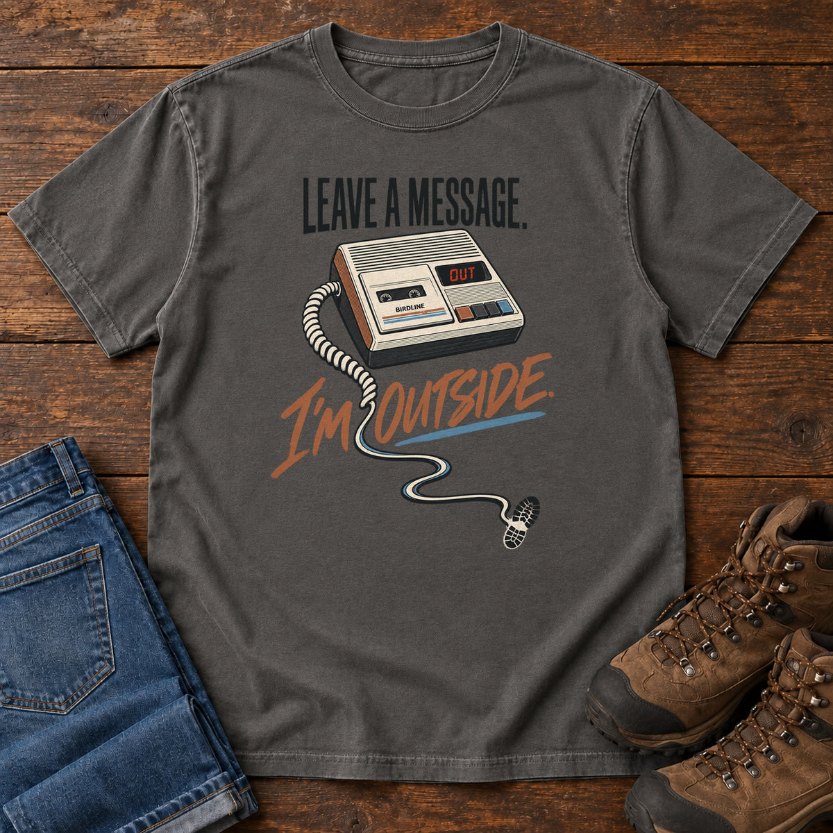 Pepper Leave a Message I'm Outside T-Shirt with a retro answering machine, trailing cord and orange script, front flat lay on wood.