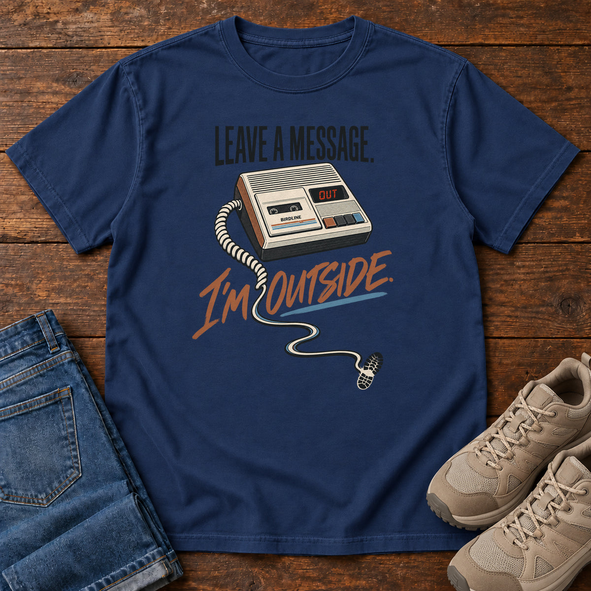 True Navy Leave a Message I'm Outside T-Shirt with a retro answering machine, trailing cord and orange script, front flat lay on wood.