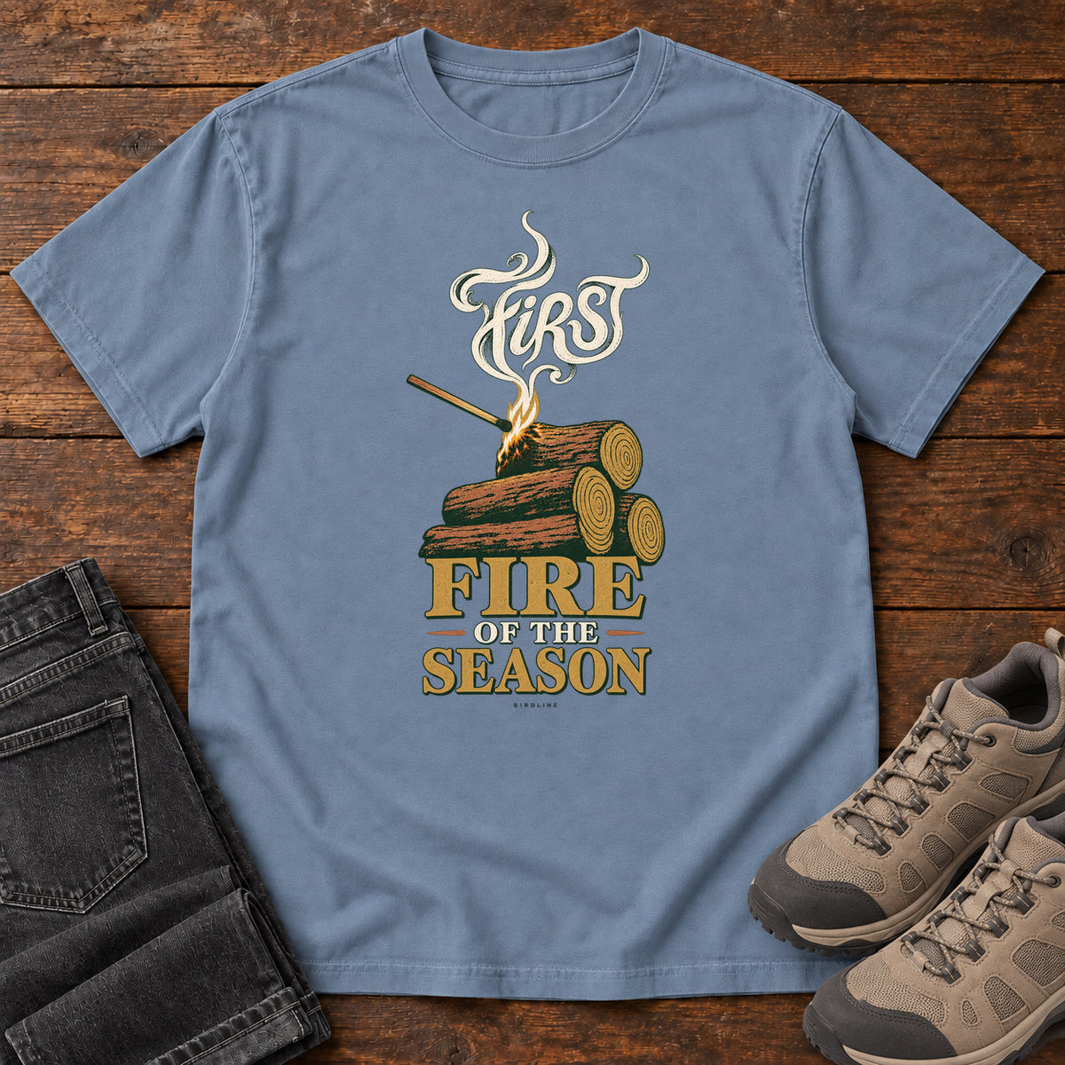 Blue Jean First Fire of the Season T-Shirt with stacked logs, a small flame and curling smoke around First Fire of the Season lettering; front flat lay on wood.