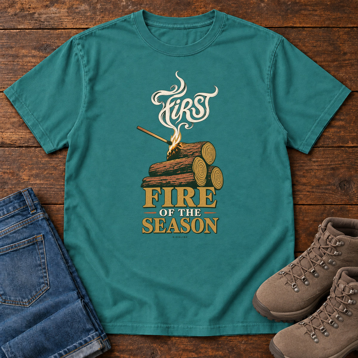 Emerald First Fire of the Season T-Shirt with stacked logs, a small flame and curling smoke around First Fire of the Season lettering; front flat lay on wood.