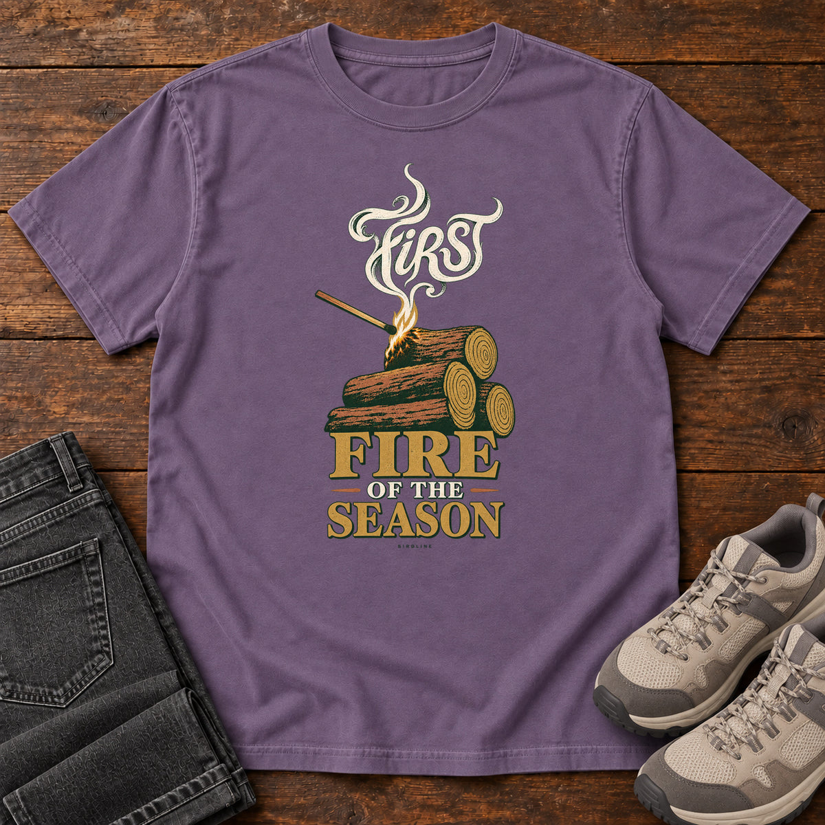 Grape First Fire of the Season T-Shirt with stacked logs, a small flame and curling smoke around First Fire of the Season lettering; front flat lay on wood.