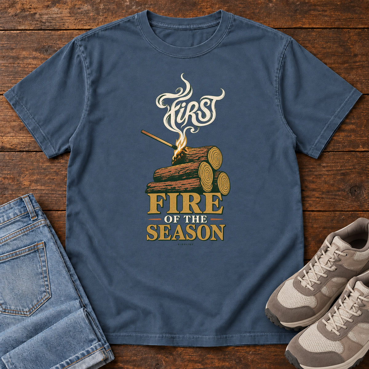 Midnight First Fire of the Season T-Shirt with stacked logs, a small flame and curling smoke around First Fire of the Season lettering; front flat lay on wood.