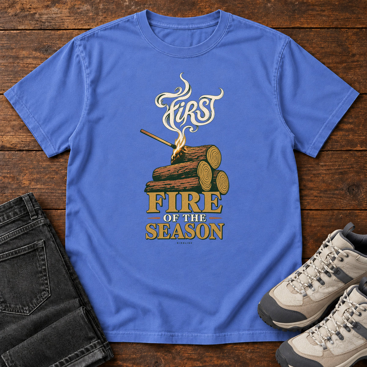 Mystic Blue First Fire of the Season T-Shirt with stacked logs, a small flame and curling smoke around First Fire of the Season lettering; front flat lay on wood.