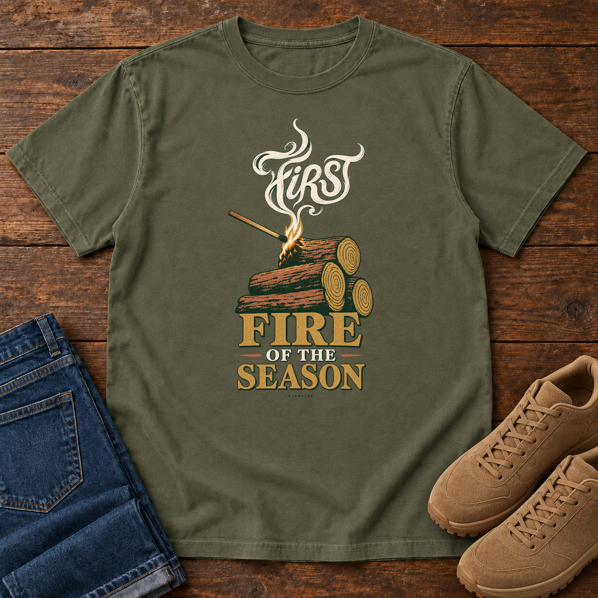 Sage First Fire of the Season T-Shirt with stacked logs, a small flame and curling smoke around First Fire of the Season lettering; front flat lay on wood.