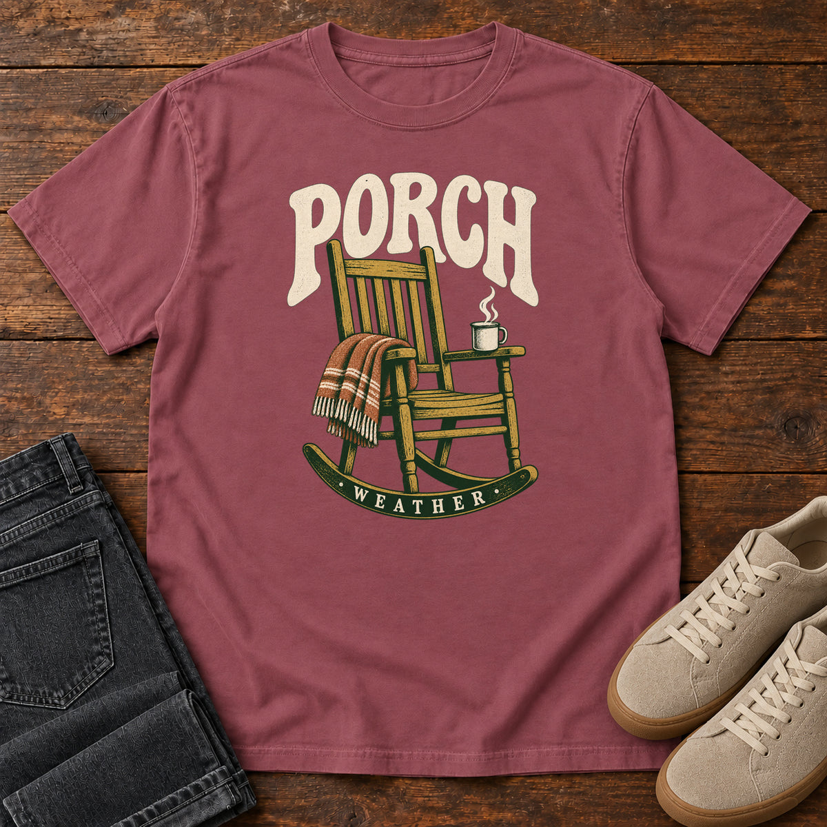 Brick Porch Weather T-Shirt with a wooden rocking chair with a striped blanket and steaming mug; front flat lay on wood.