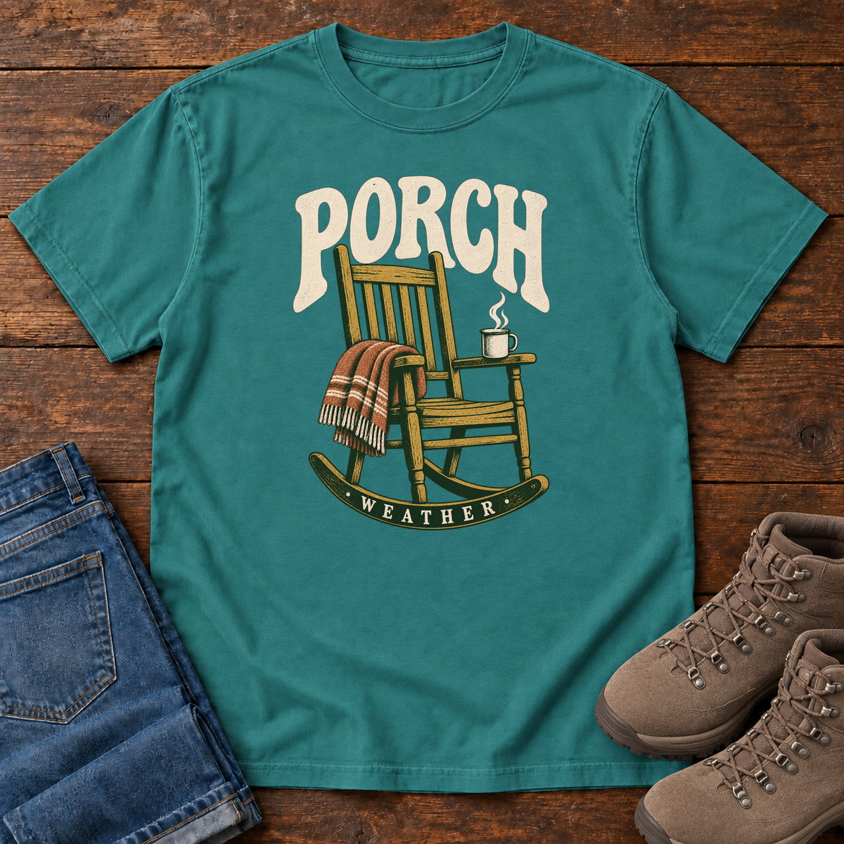 Emerald Porch Weather T-Shirt with a wooden rocking chair with a striped blanket and steaming mug; front flat lay on wood.