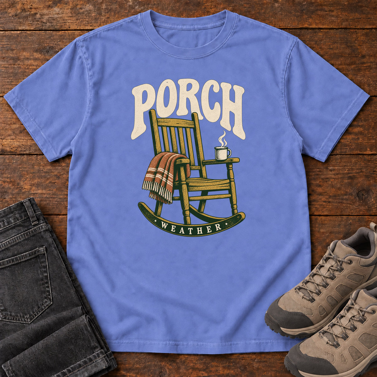 Flo Blue Porch Weather T-Shirt with a wooden rocking chair with a striped blanket and steaming mug; front flat lay on wood.