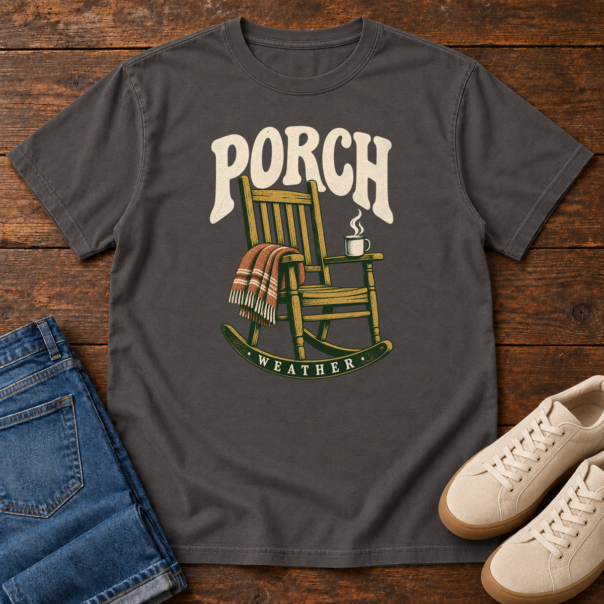 Graphite Porch Weather T-Shirt with a wooden rocking chair with a striped blanket and steaming mug; front flat lay on wood.
