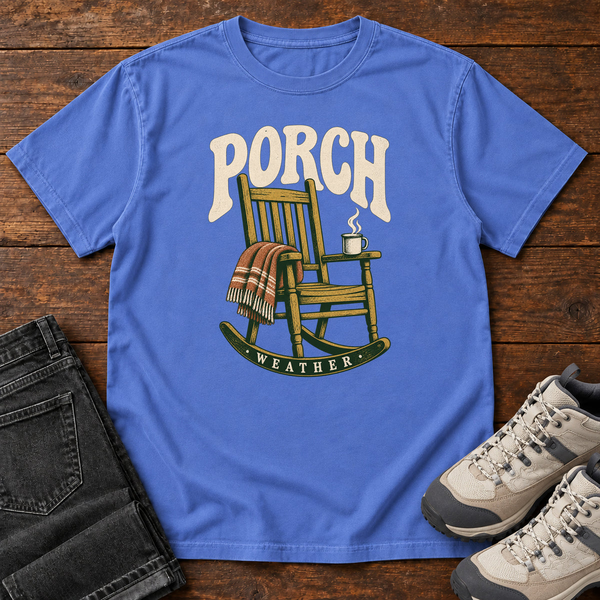 Mystic Blue Porch Weather T-Shirt with a wooden rocking chair with a striped blanket and steaming mug; front flat lay on wood.