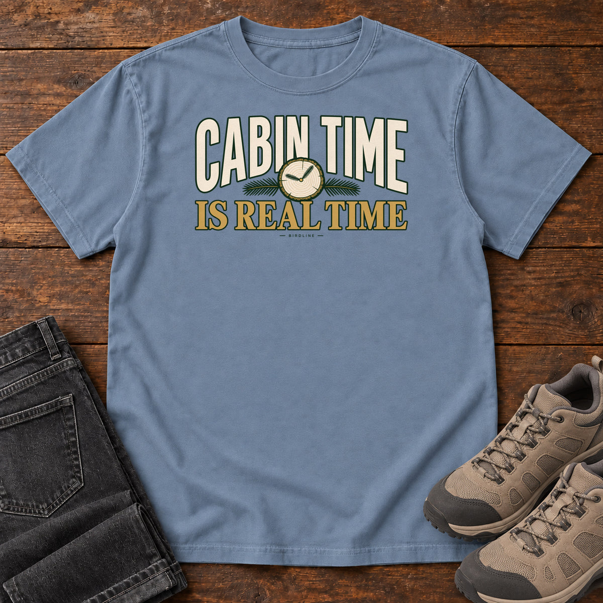 Blue Jean Cabin Time Is Real Time T-Shirt with cream and gold lettering surrounding a small clock illustration, front flat lay on wood.