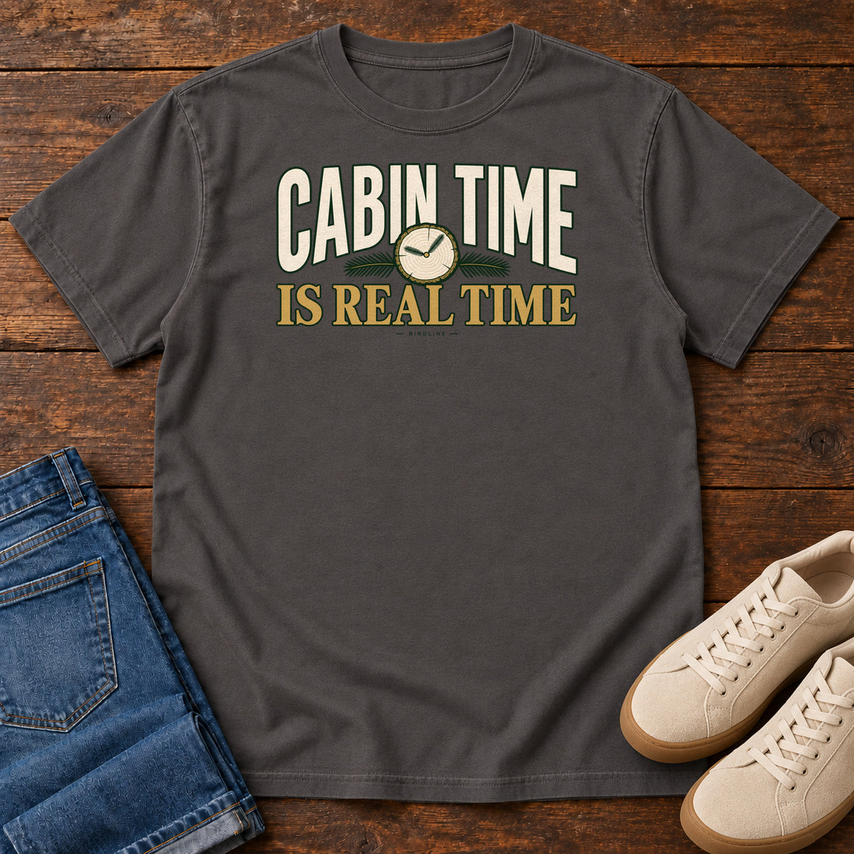 Graphite Cabin Time Is Real Time T-Shirt with cream and gold lettering surrounding a small clock illustration, front flat lay on wood.