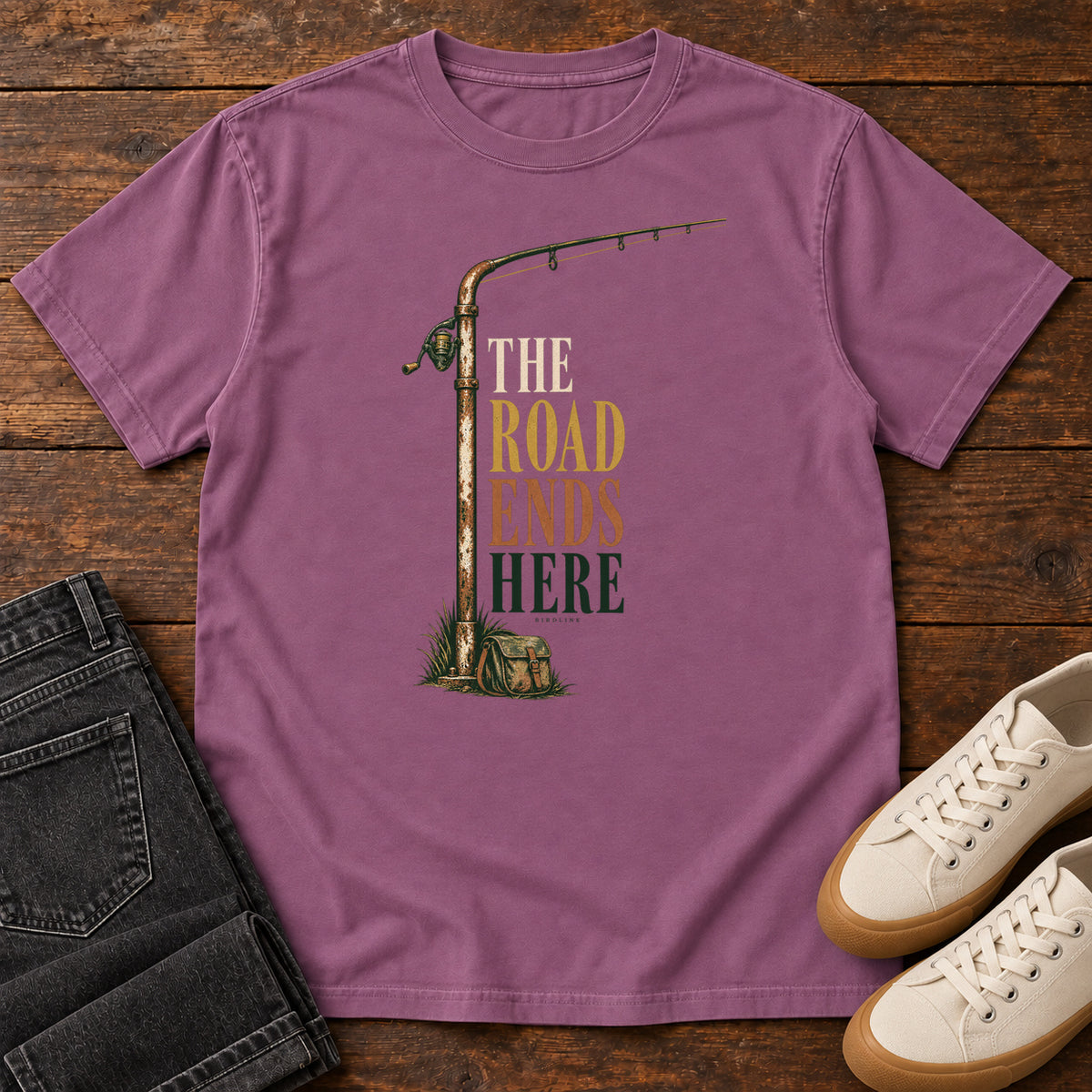 The Road Ends Here T-Shirt in Berry, front laid flat on wood, featuring a fishing rod beside stacked earth-tone slogan lettering.