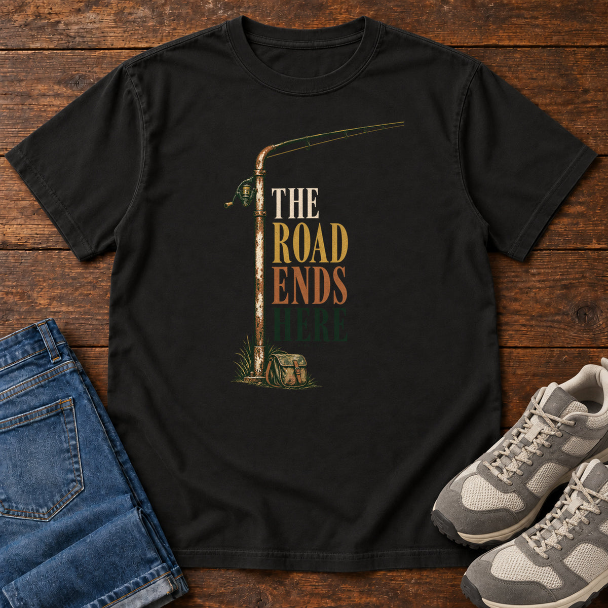 The Road Ends Here T-Shirt in Black, front laid flat on wood, featuring a fishing rod beside stacked earth-tone slogan lettering.