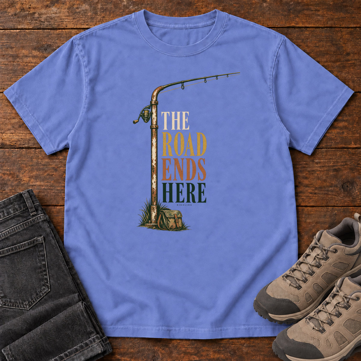 The Road Ends Here T-Shirt in Flo Blue, front laid flat on wood, featuring a fishing rod beside stacked earth-tone slogan lettering.