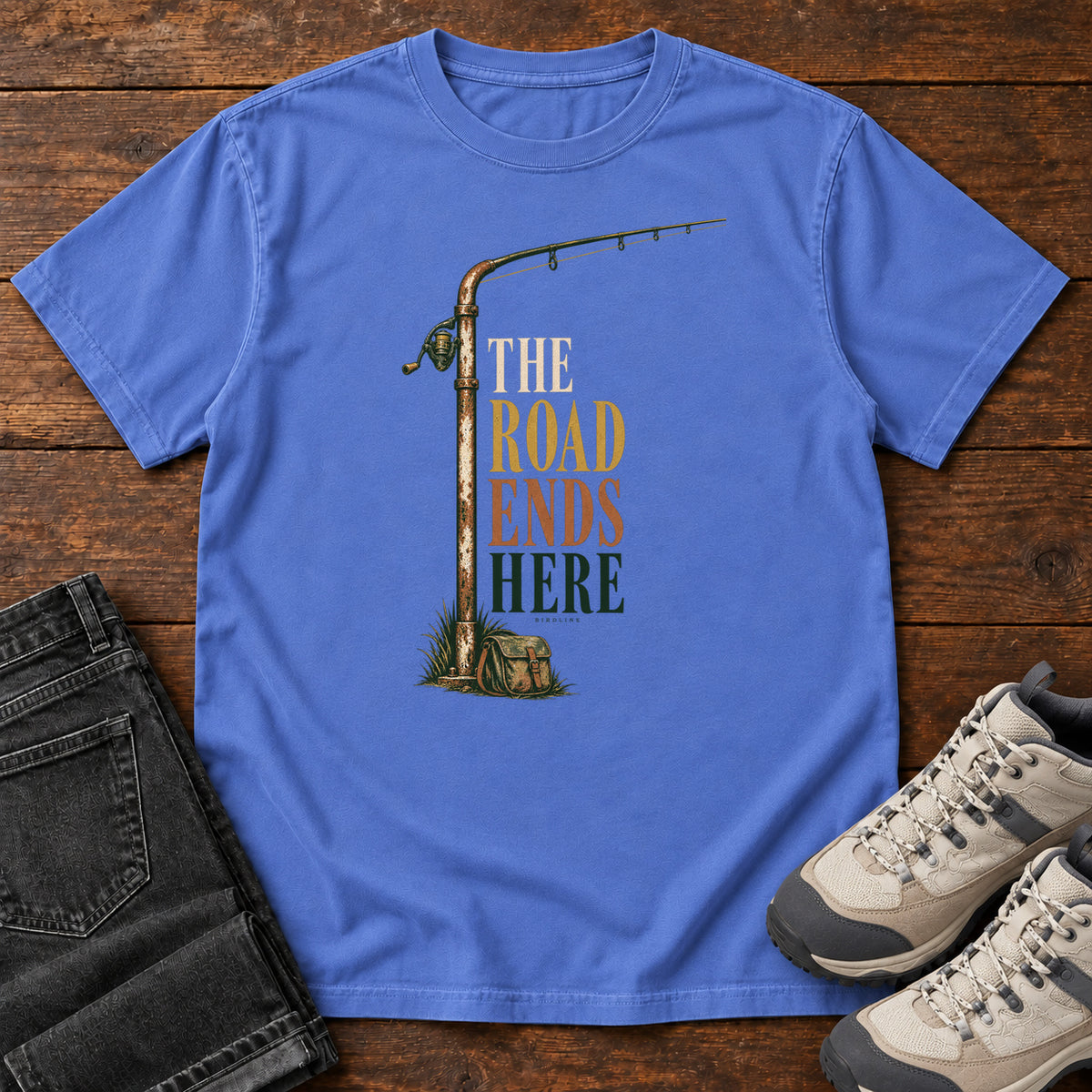 The Road Ends Here T-Shirt in Mystic Blue, front laid flat on wood, featuring a fishing rod beside stacked earth-tone slogan lettering.