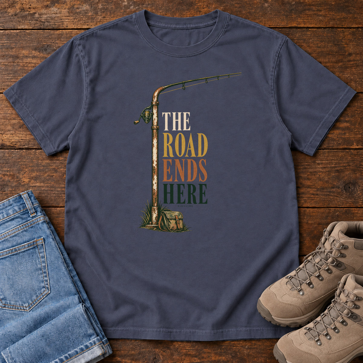 The Road Ends Here T-Shirt in Navy, front laid flat on wood, featuring a fishing rod beside stacked earth-tone slogan lettering.